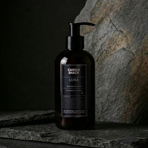 Black bottle hand and body wash labeled 'Candle Shack Luna' on a dark stone surface with a stone wall background